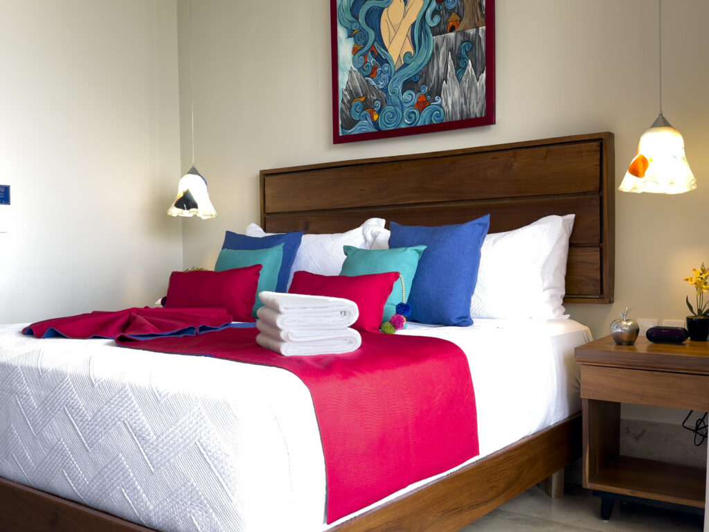Wooden bed with white bedding, a red throw, colorful pillows, and folded towels beneath pendant lights.
