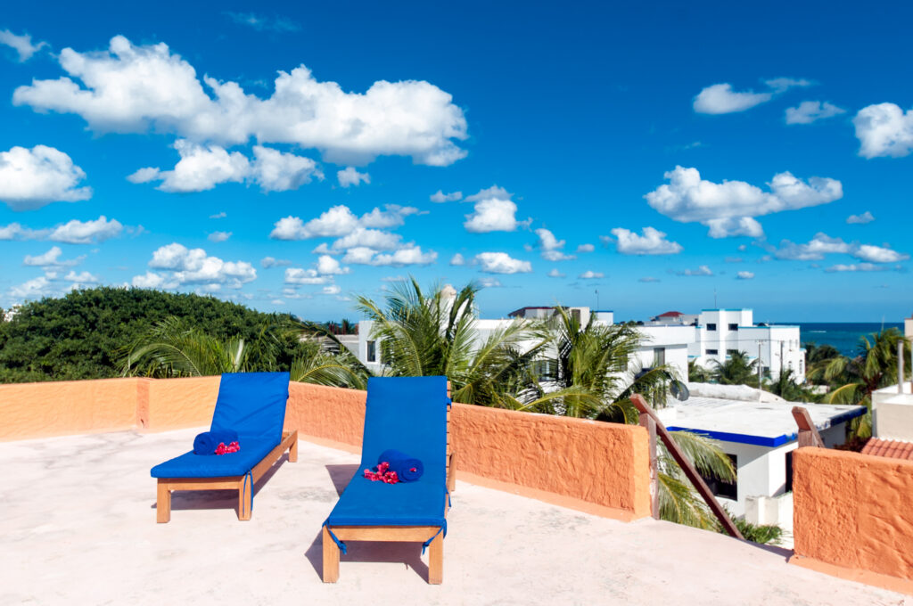 Two blue lounge chairs on a rooftop terrace overlooking treetops, buildings, and the distant sea.