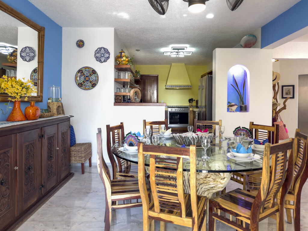 Dining table and sideboard beside decorative wall plates, with a yellow-countered kitchen beyond.