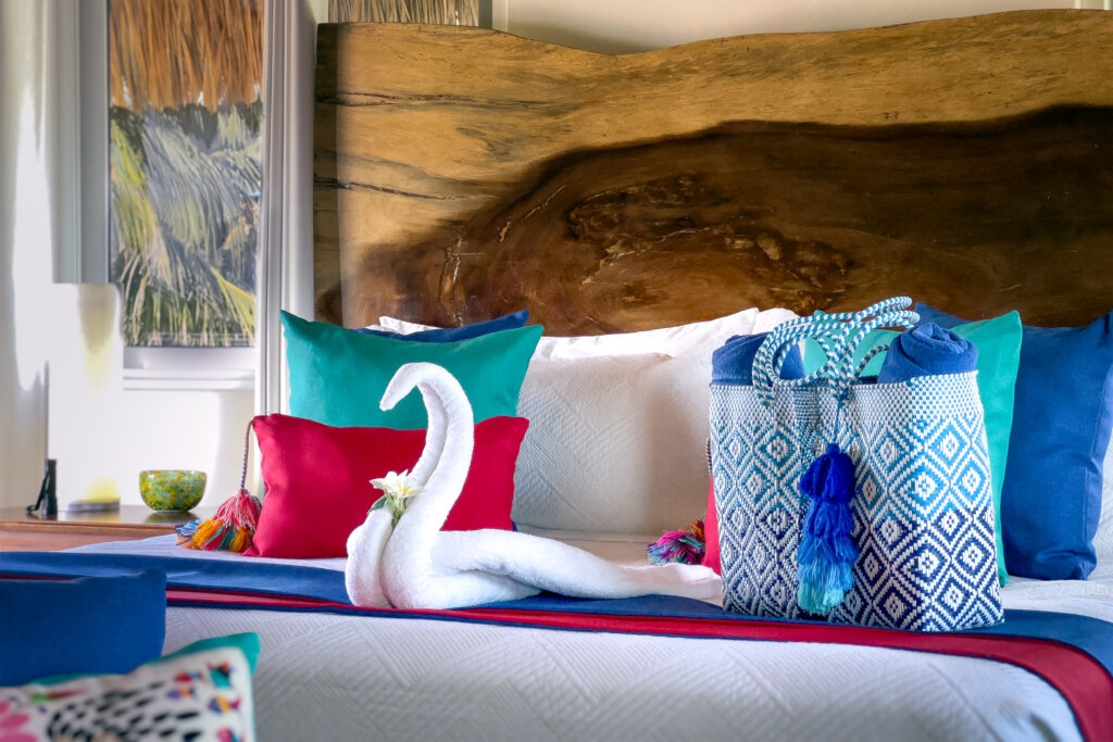 Towel swan and woven blue bag on a bed with bright pillows and a natural wood headboard.