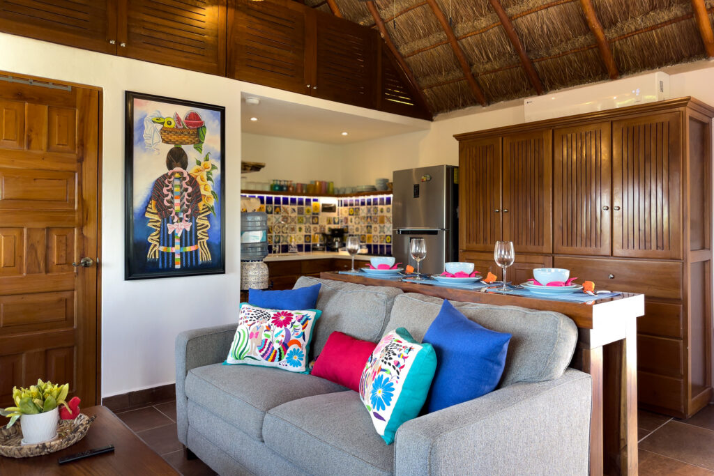 Gray sofa with colorful cushions in front of the kitchen, wooden cabinets, and framed artwork.