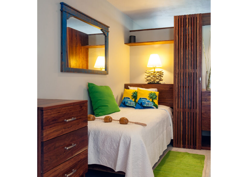 Single bed with green and yellow cushions beside a dresser, mirror, and wooden wardrobe.