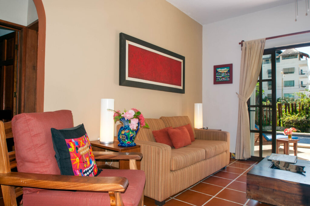 Sofa with orange cushions and a wooden armchair beside a lamp and vase of flowers.
