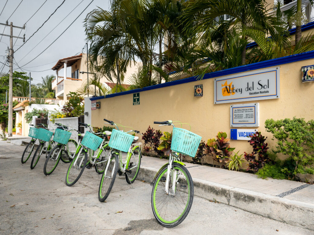 Bicycles with turquoise baskets lined up beside the Abbey del Sol sign and entrance wall.