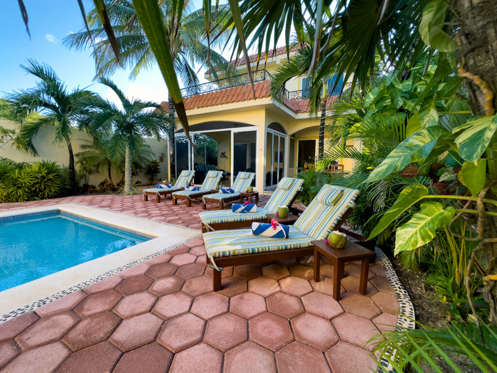 Pool and lounge chairs among dense tropical plants beside a yellow two-story house.