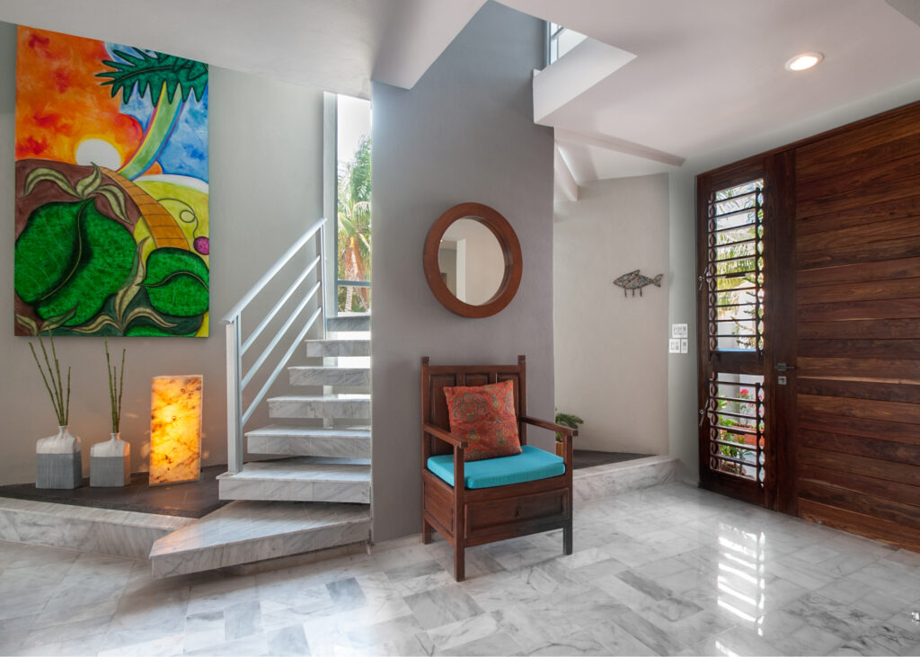 Wooden chair with a turquoise cushion beside the front door, staircase, and colorful artwork.
