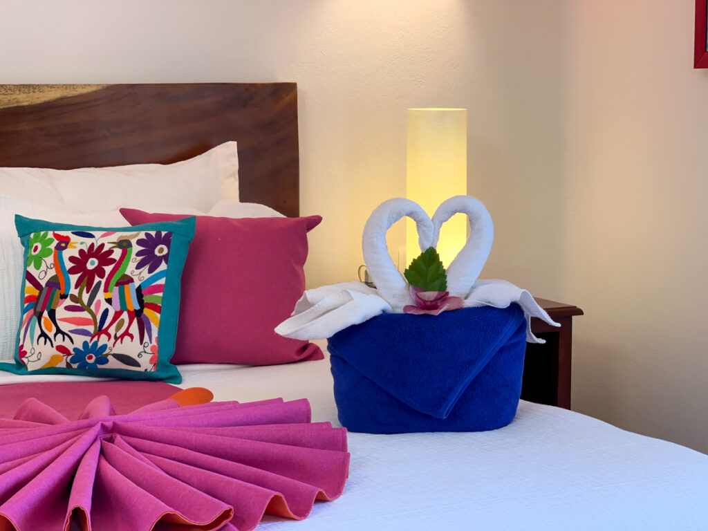 White towel swans on a blue towel beside a fan-folded pink throw and colorful pillows.