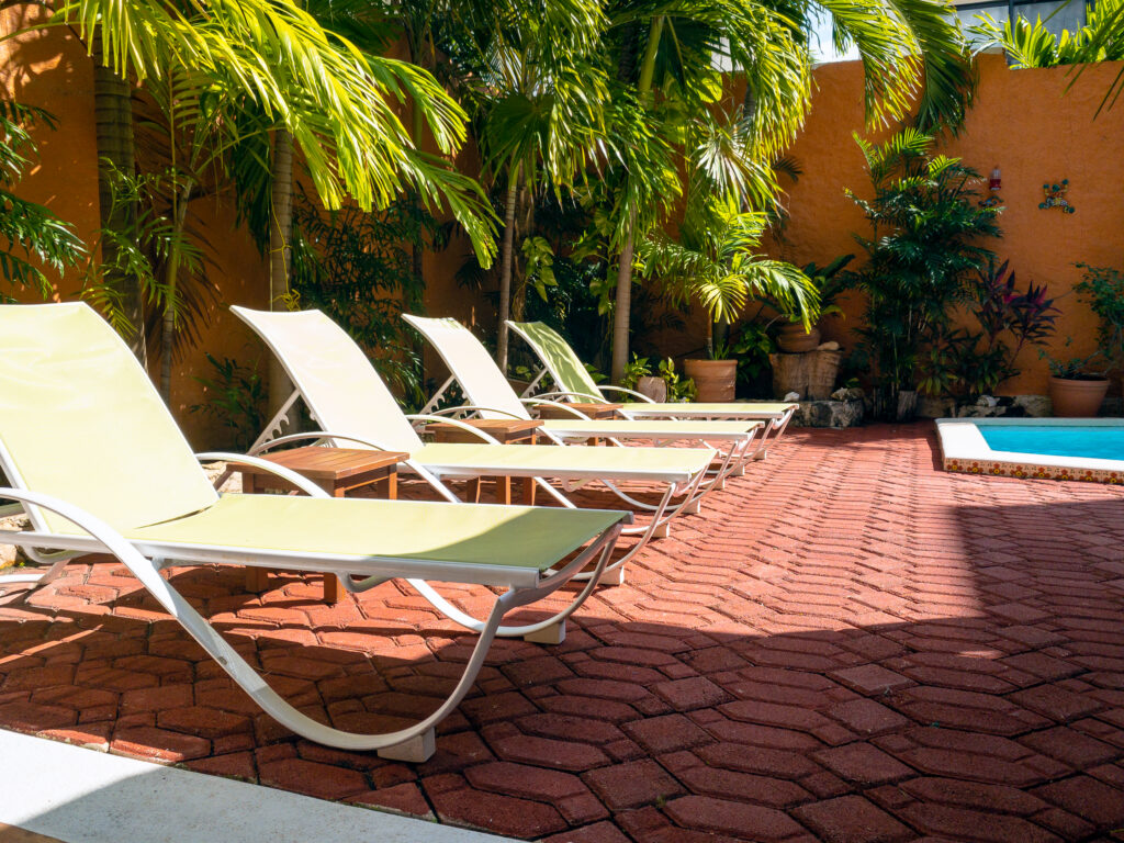 Light-colored lounge chairs on a red-paved patio beside palms and the edge of a pool.