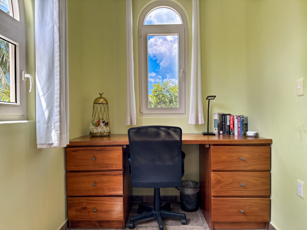 Wooden desk with drawers, a chair, lamp, and books beneath a narrow arched window.