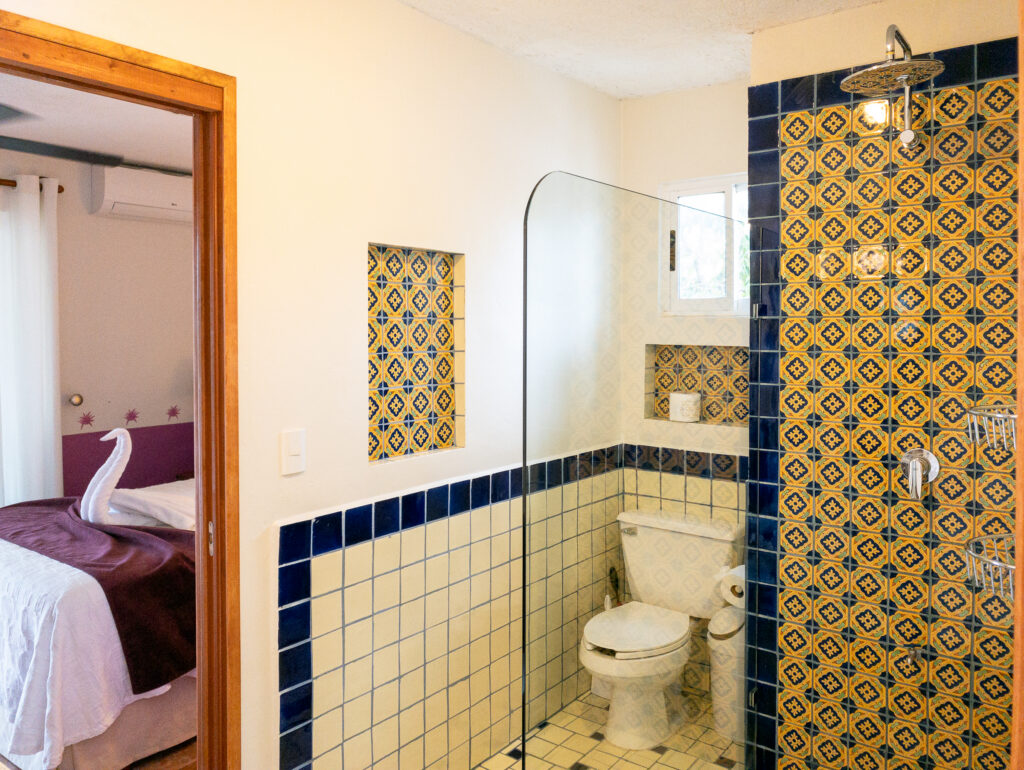 Shower with patterned yellow-and-blue tiles beside a toilet and bedroom doorway.