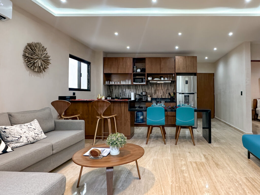 Gray sofa and wooden coffee table facing a kitchen with wooden cabinets, barstools, and turquoise dining chairs.