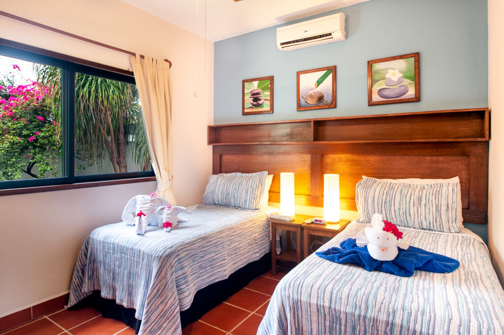 Two single beds with striped covers beside a large window and wooden headboard wall.