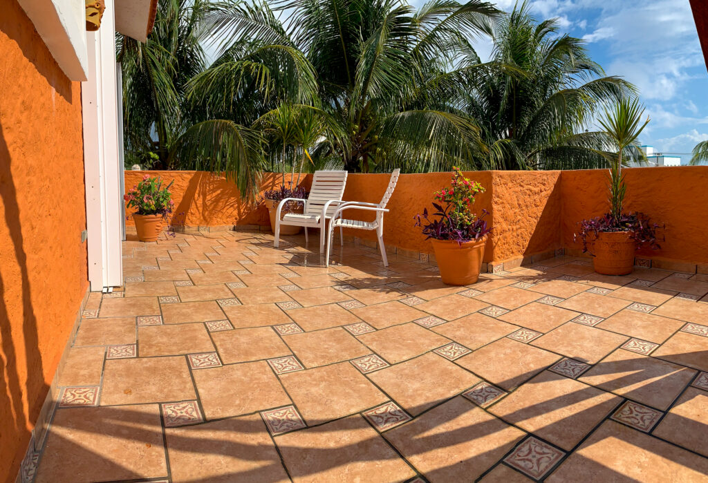 Broad tiled terrace with a white chair, potted plants, and orange walls overlooking palms.