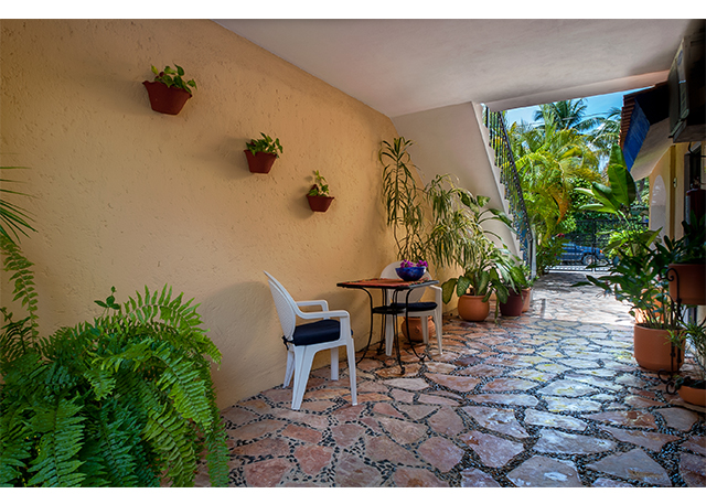 Small table and chairs along a covered stone passage lined with potted tropical plants.