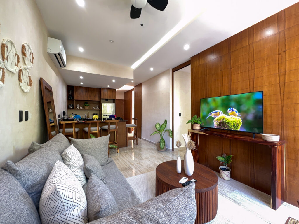 Gray sofa and round coffee table facing a television on a wood-paneled wall, with kitchen beyond.