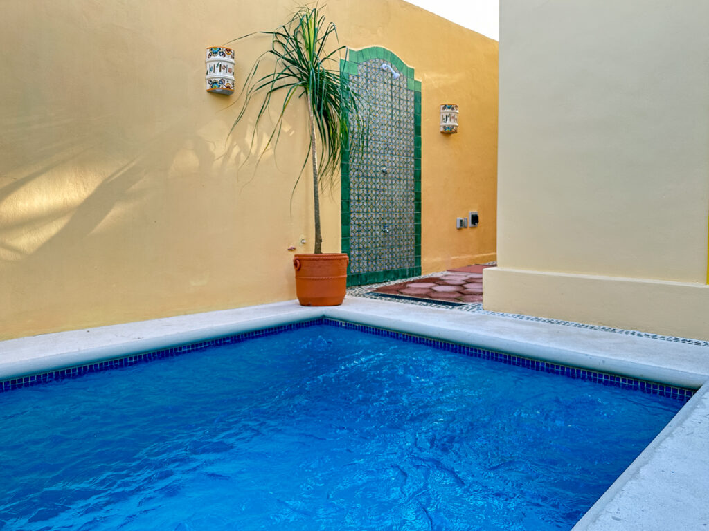 Blue pool beside a green-tiled outdoor shower and a potted palm against a yellow wall.