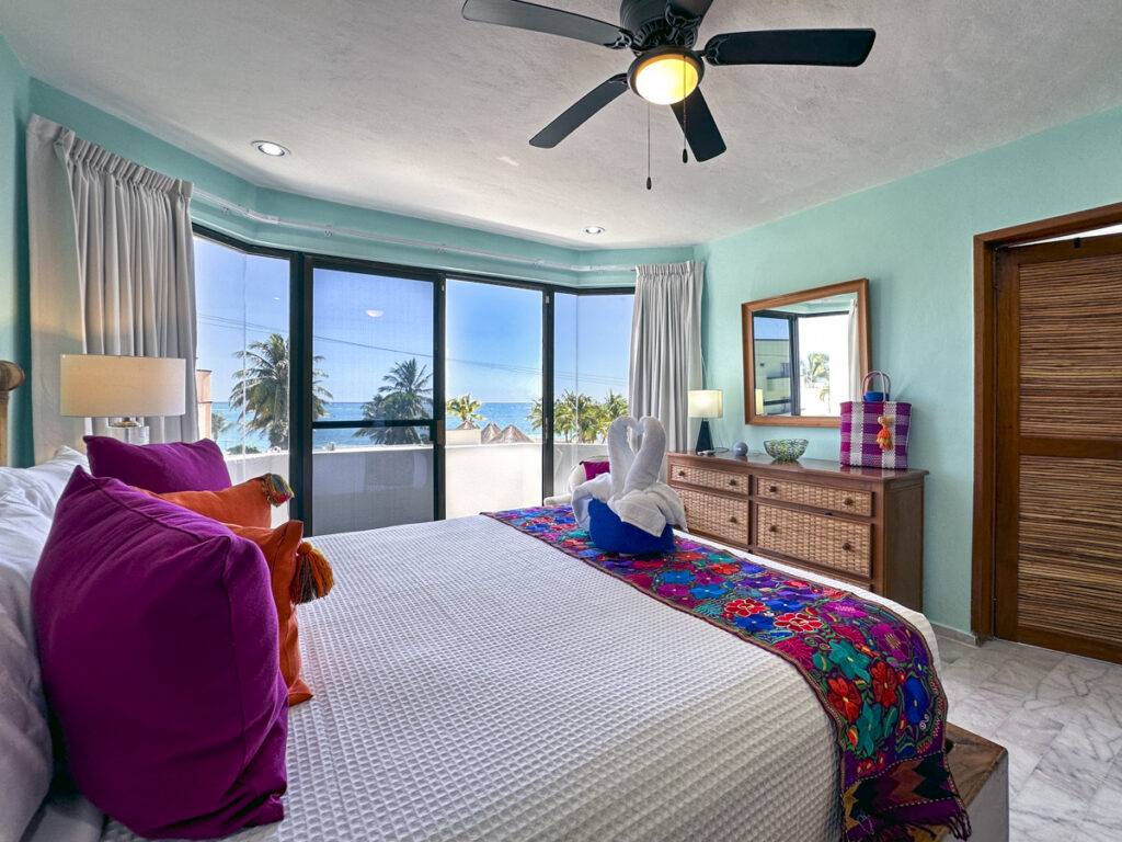Bed with a floral runner faces broad windows overlooking palms and the sea.