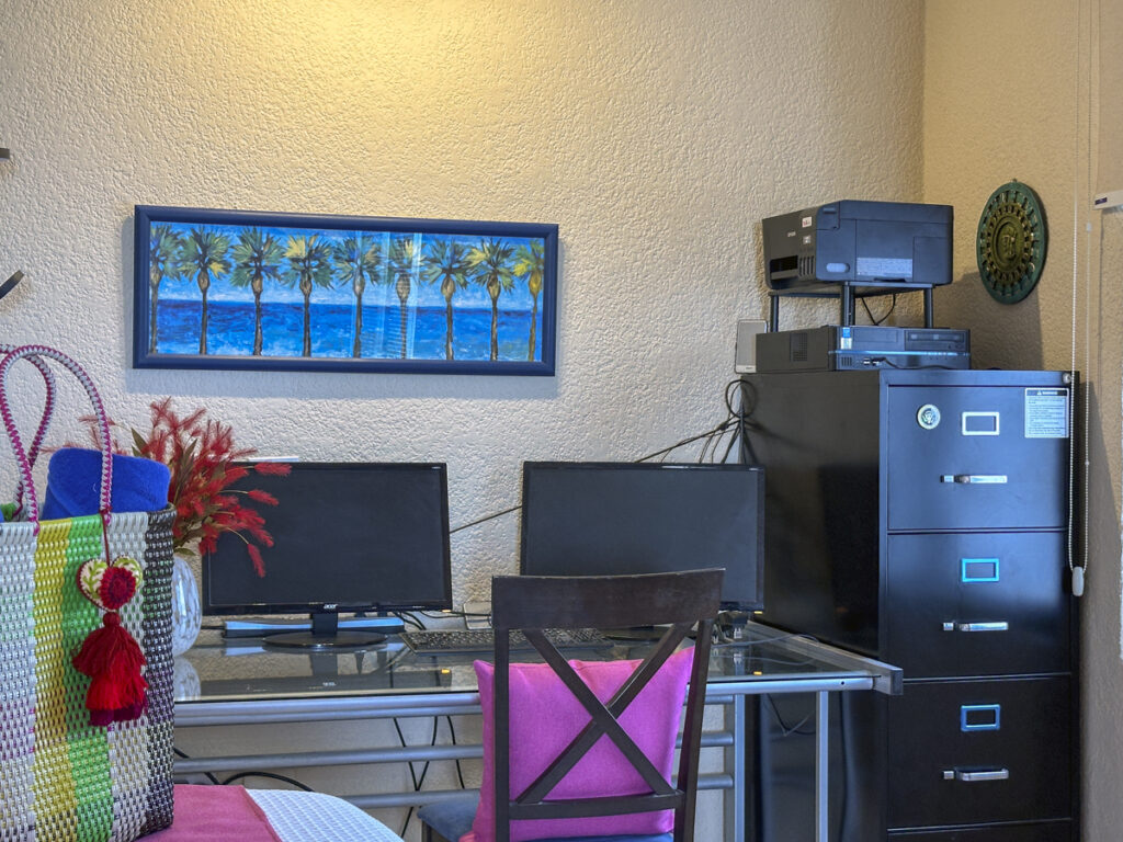 Desk with a monitor and chair beside a filing cabinet and printer beneath blue artwork.