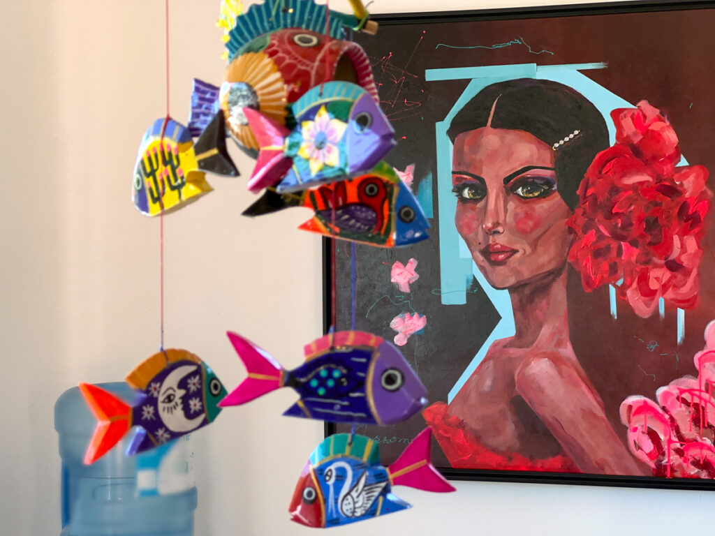 Colorful hanging fish ornaments in front of a portrait with red flowers.