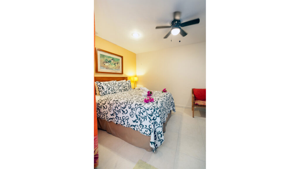 Bedroom with a patterned bedspread, ceiling fan, and a wicker chair beside the bed.