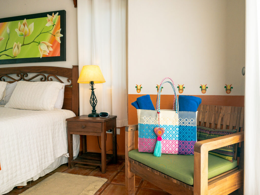 Colorful woven bag on a wooden chair beside a bed and bedside lamp.