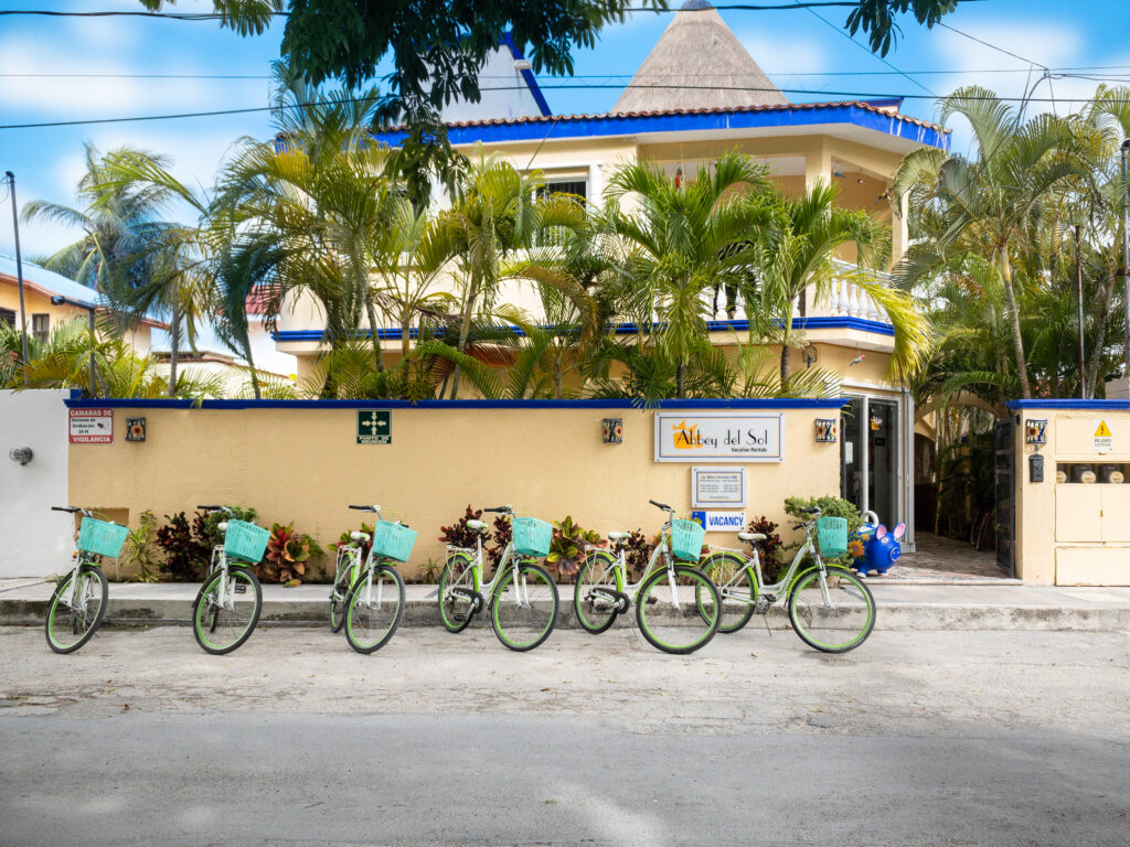 Bicycles lined up outside the yellow Abbey del Sol building beneath palm trees.