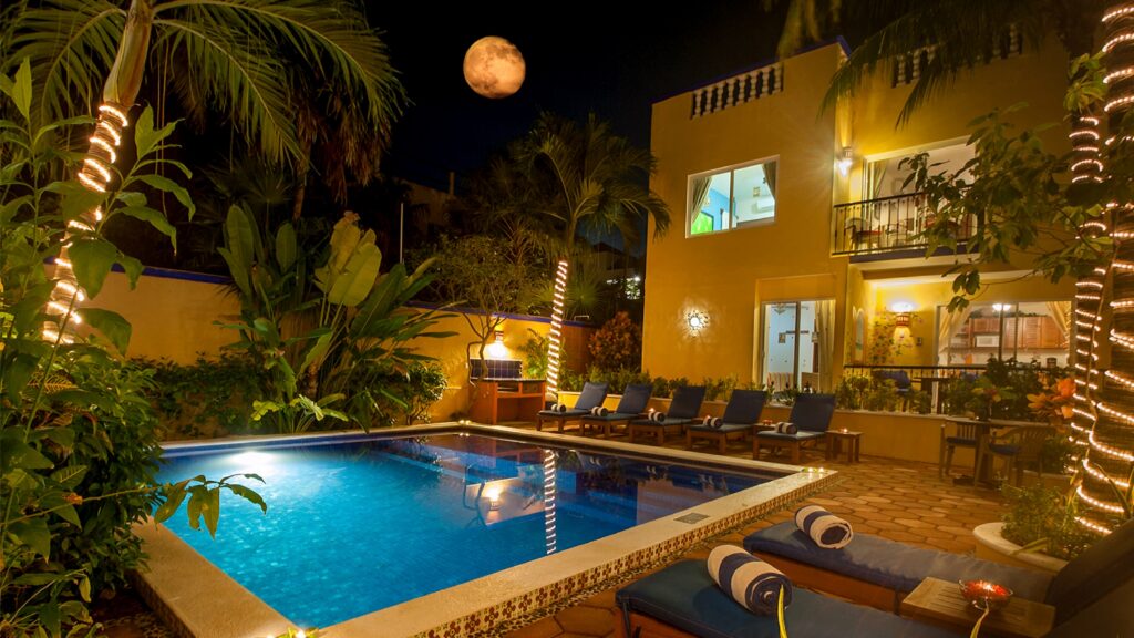 Illuminated pool and lounge chairs beneath palm trees beside a yellow building at night.
