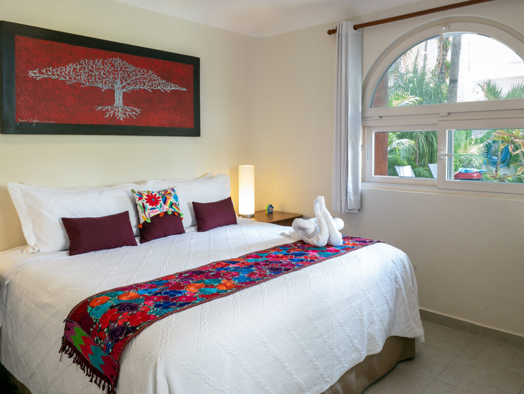 White bed with a floral runner and dark cushions beneath red artwork beside an arched window.