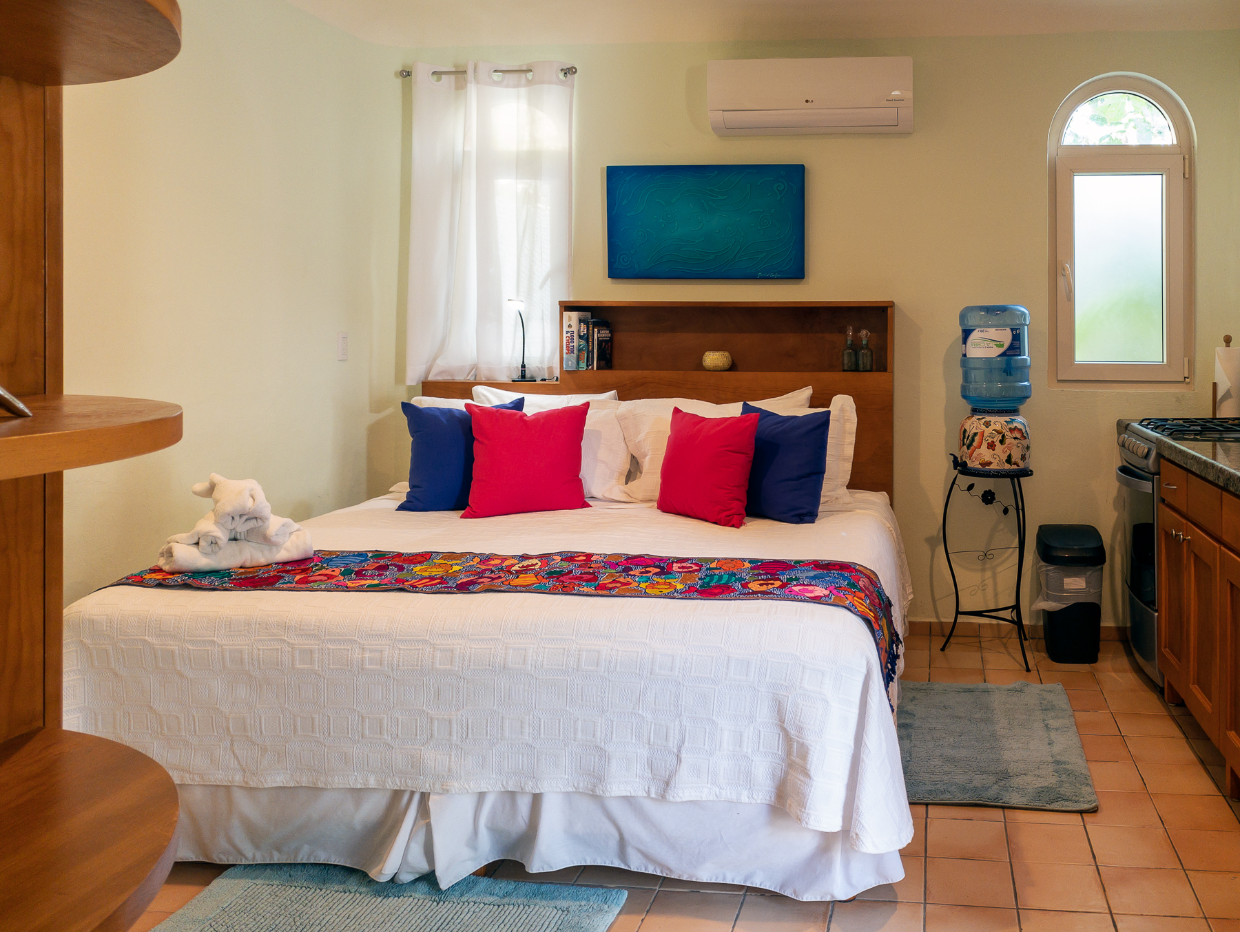White bed with red and blue cushions and a floral runner beneath blue artwork.