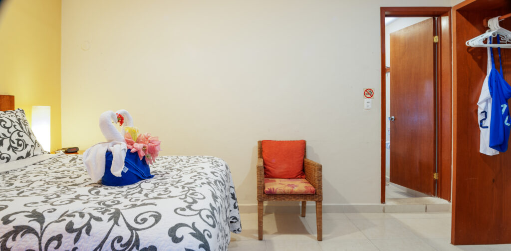 Bed with a folded towel decoration beside a wicker chair and open wooden storage.