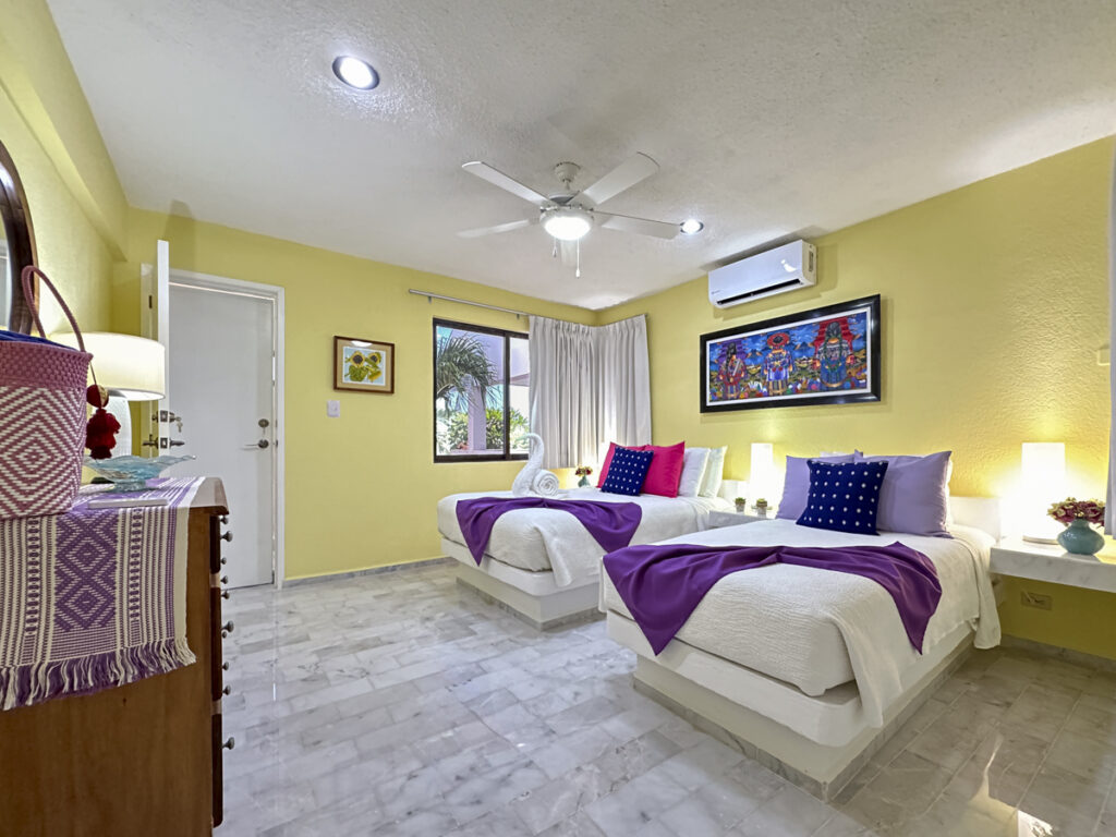 Two beds with purple throws in a yellow-walled room with a dresser, mirror, and ceiling fan.