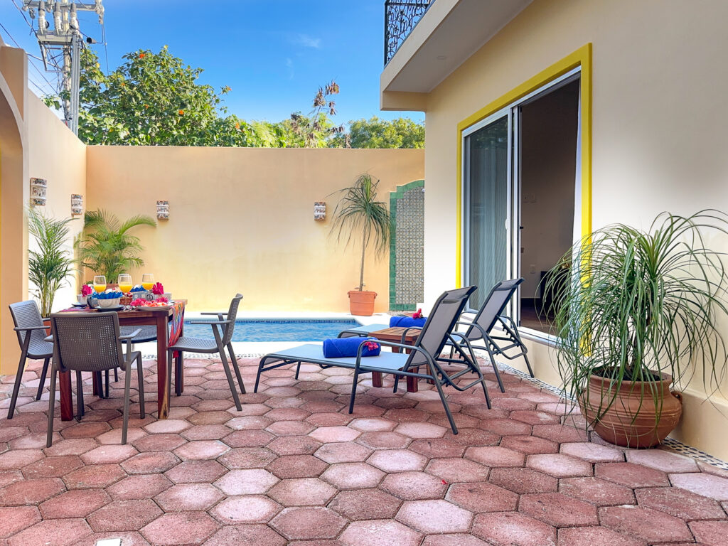 Lounge chairs and a dining table on a stone courtyard beside the pool and potted palms.