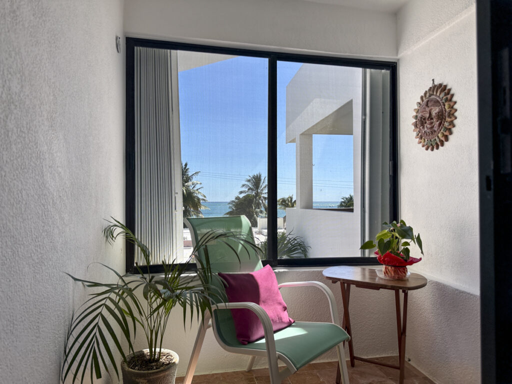 Green chair with a pink cushion beside a small table and broad window overlooking palms.