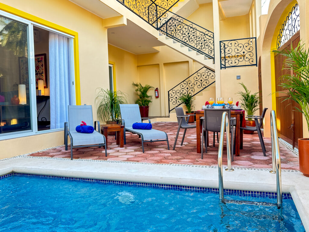Blue pool beside lounge chairs, a dining table, and an exterior staircase with decorative iron railings.