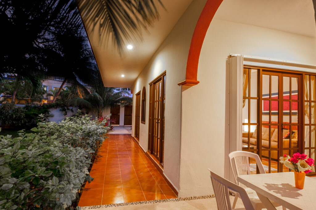 Illuminated covered walkway along the house beside garden shrubs and an outdoor table.