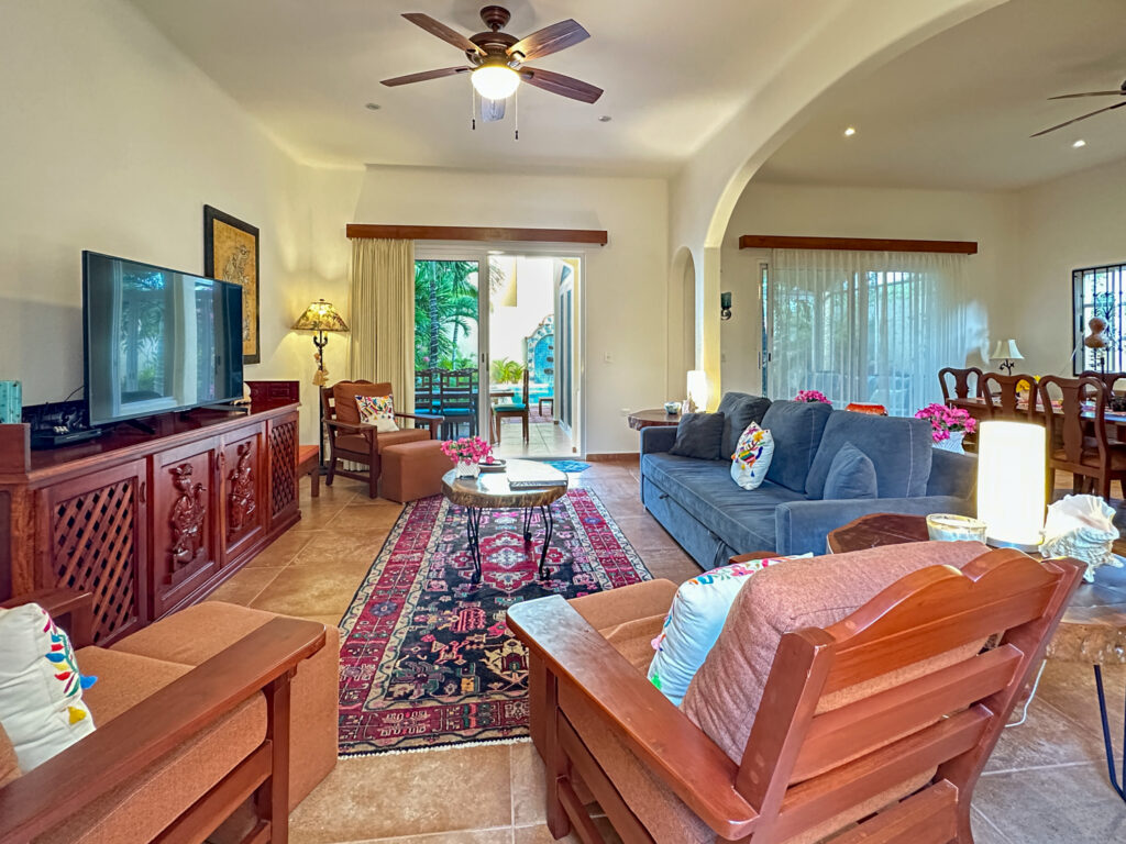 Blue sofa and wooden armchairs beside a television console, patterned rug, and garden doors.