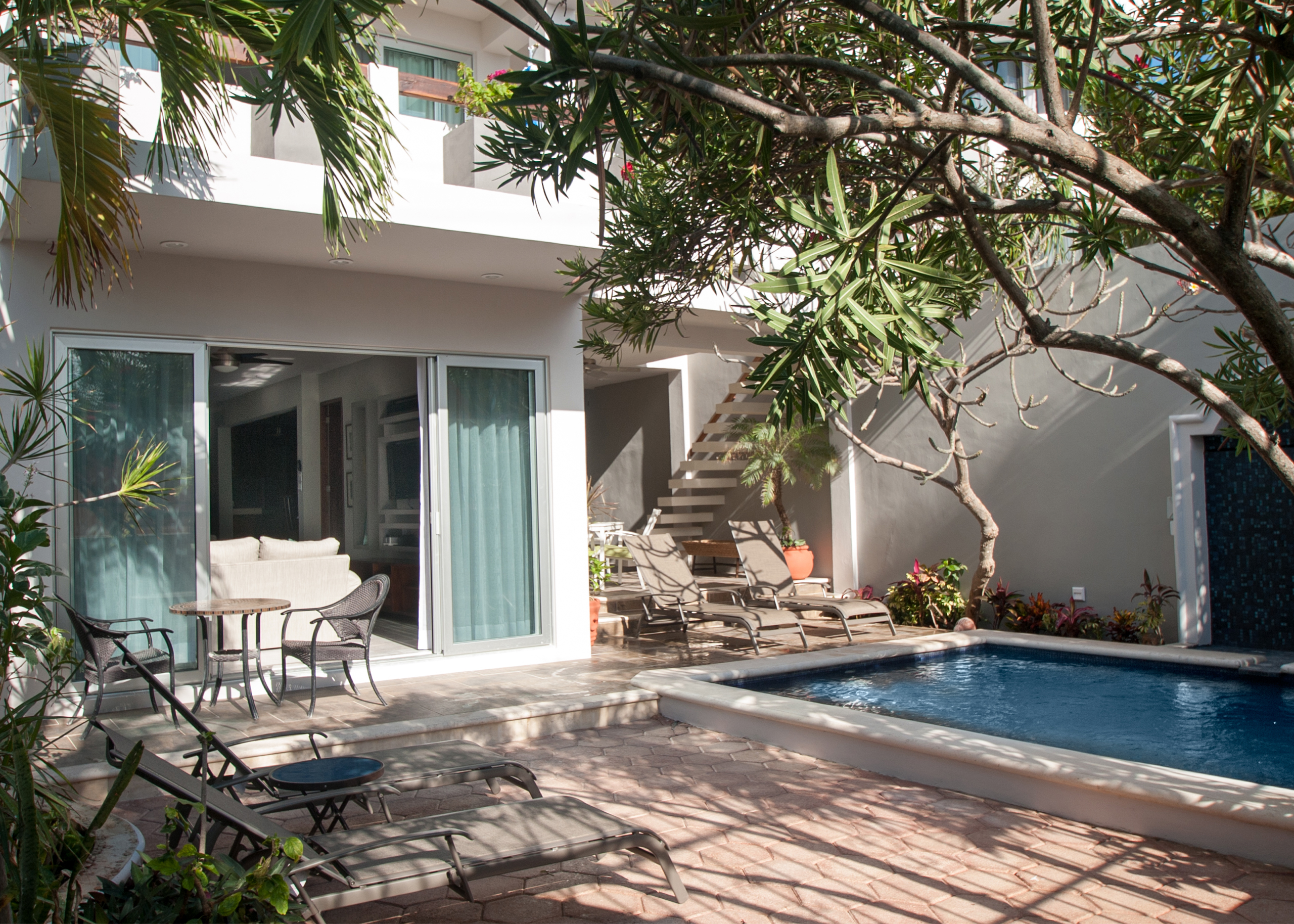 Pool beside outdoor seating and an exterior staircase beneath leafy trees.