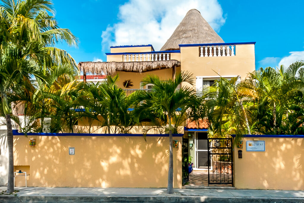 Yellow building with blue trim and a thatched rooftop structure behind palms and a gated entrance.