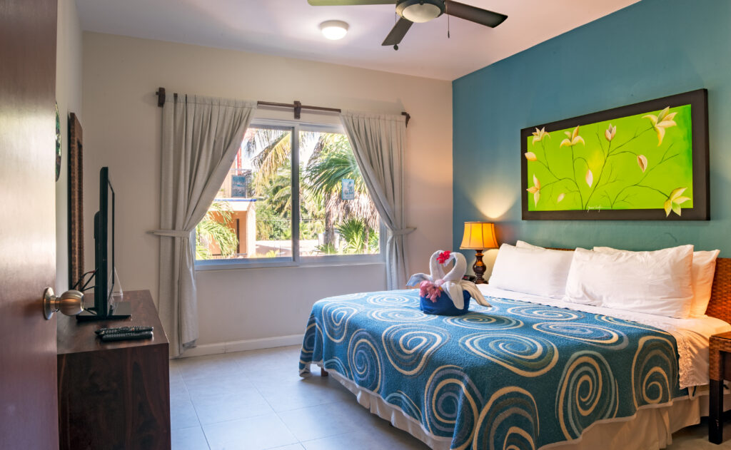 Bed with a blue spiral-patterned cover beneath green artwork beside a window overlooking palms.