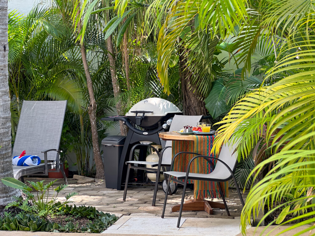 Grill beside a small table and outdoor chairs among dense palm fronds.