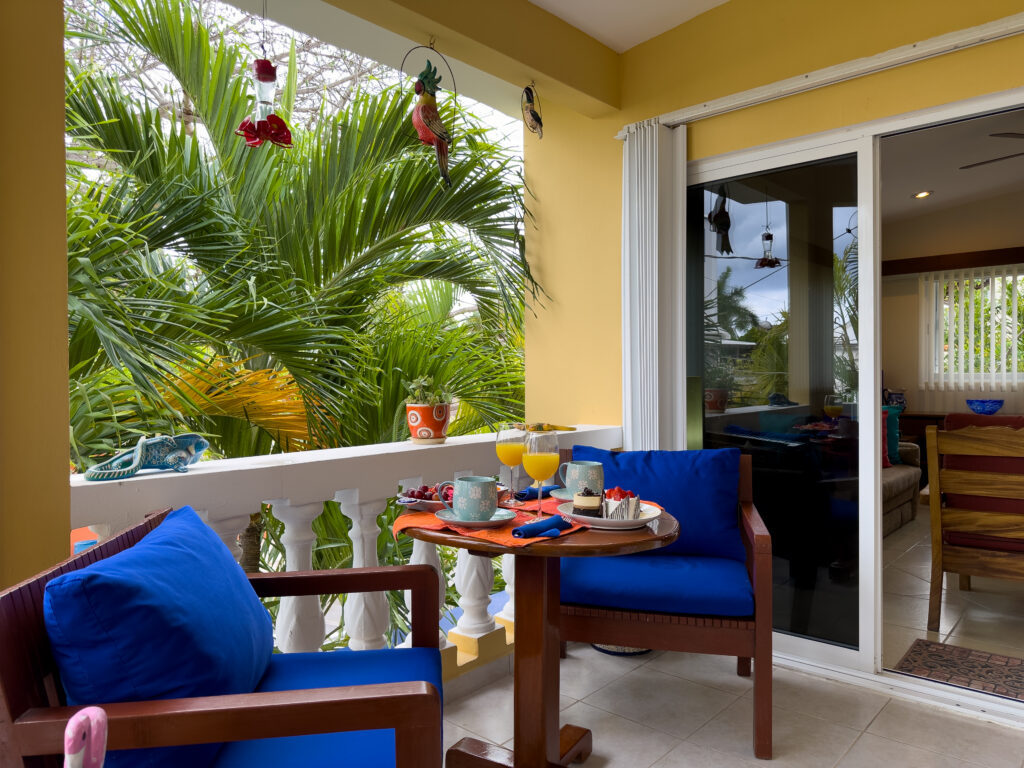 Balcony chairs with blue cushions beside a small table set with drinks and dishes among palms.