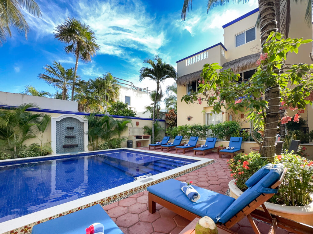 Blue-tiled pool bordered by blue lounge chairs, palms, and yellow buildings with blue trim.