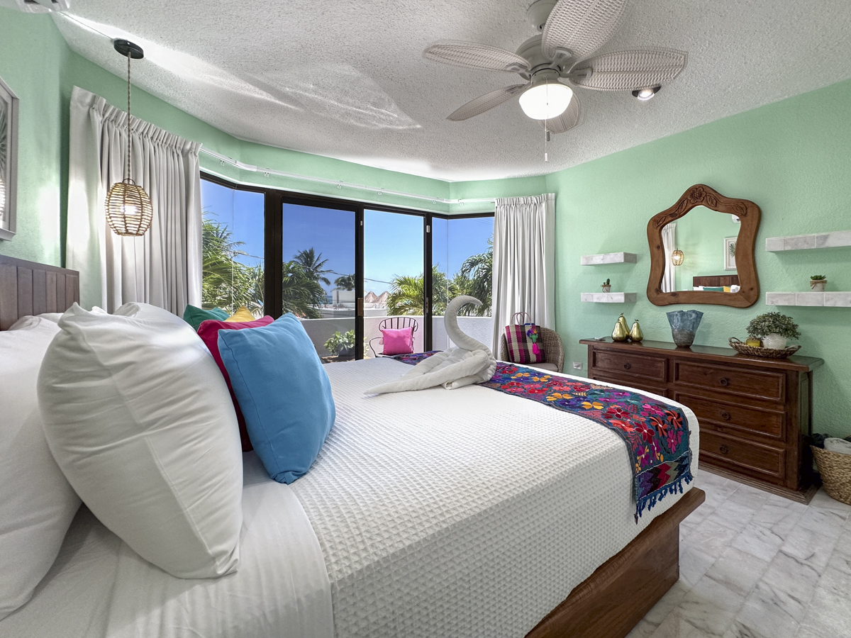 Bed with white bedding faces a wooden dresser and wide balcony doors overlooking palms.