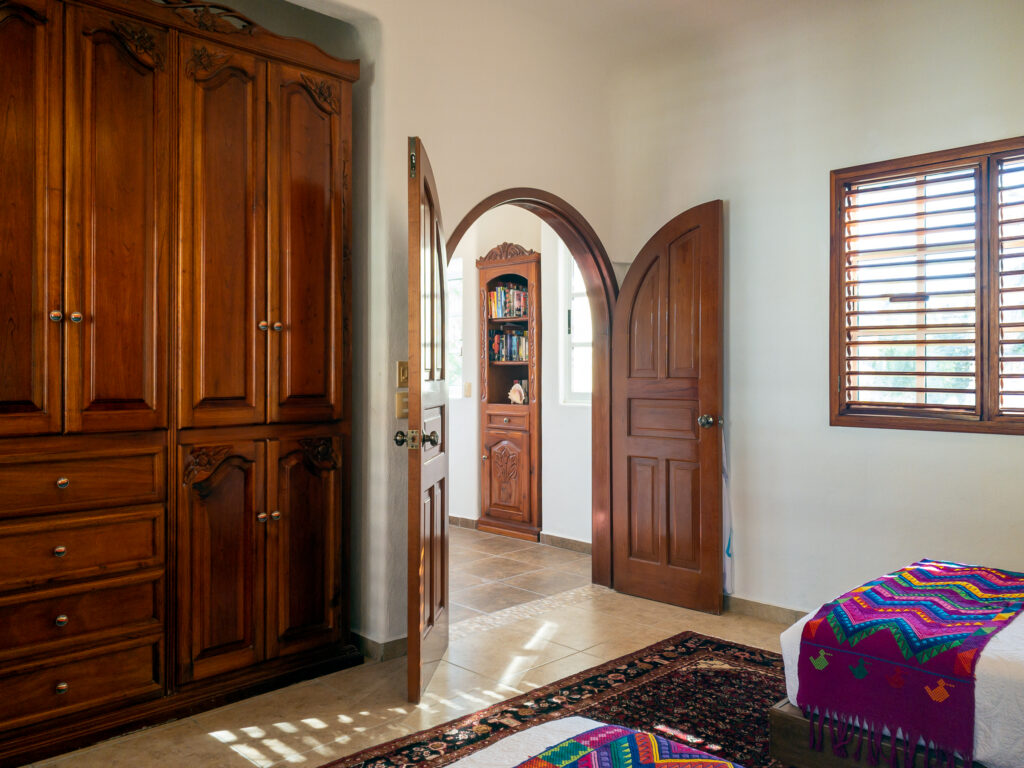 Tall carved wooden wardrobe beside an arched door and shuttered window in a bedroom.