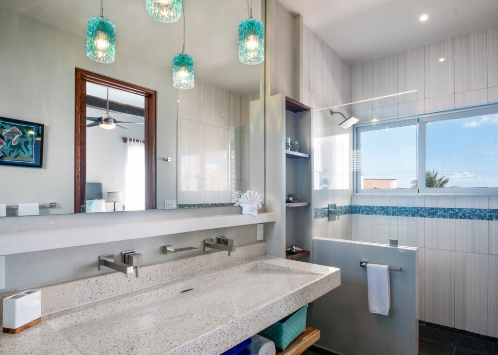 Long stone vanity beneath turquoise pendant lights beside a glass shower and wide window.