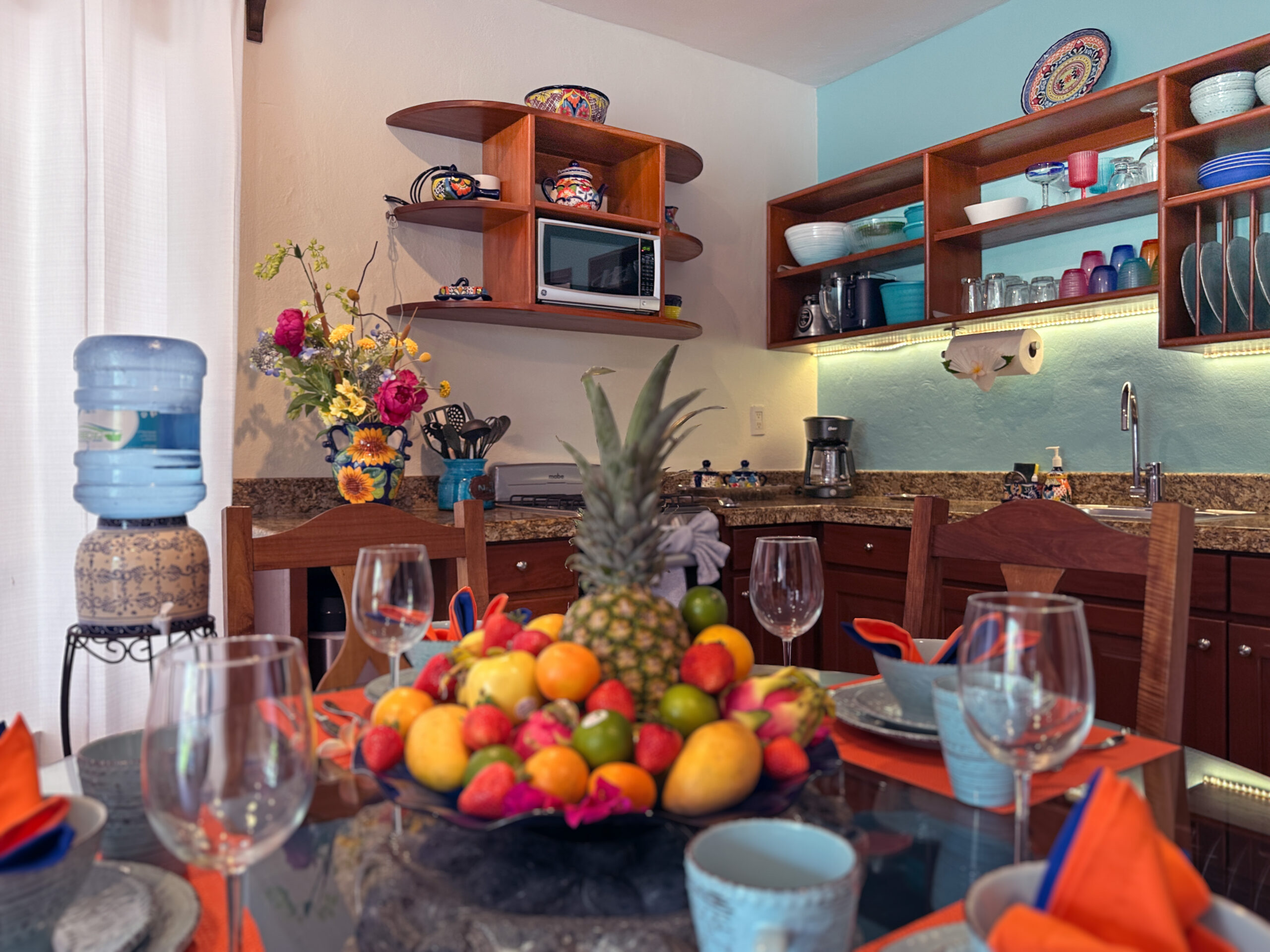 Dining table with fruit and dishes in front of wooden kitchen shelves, sink, and microwave.