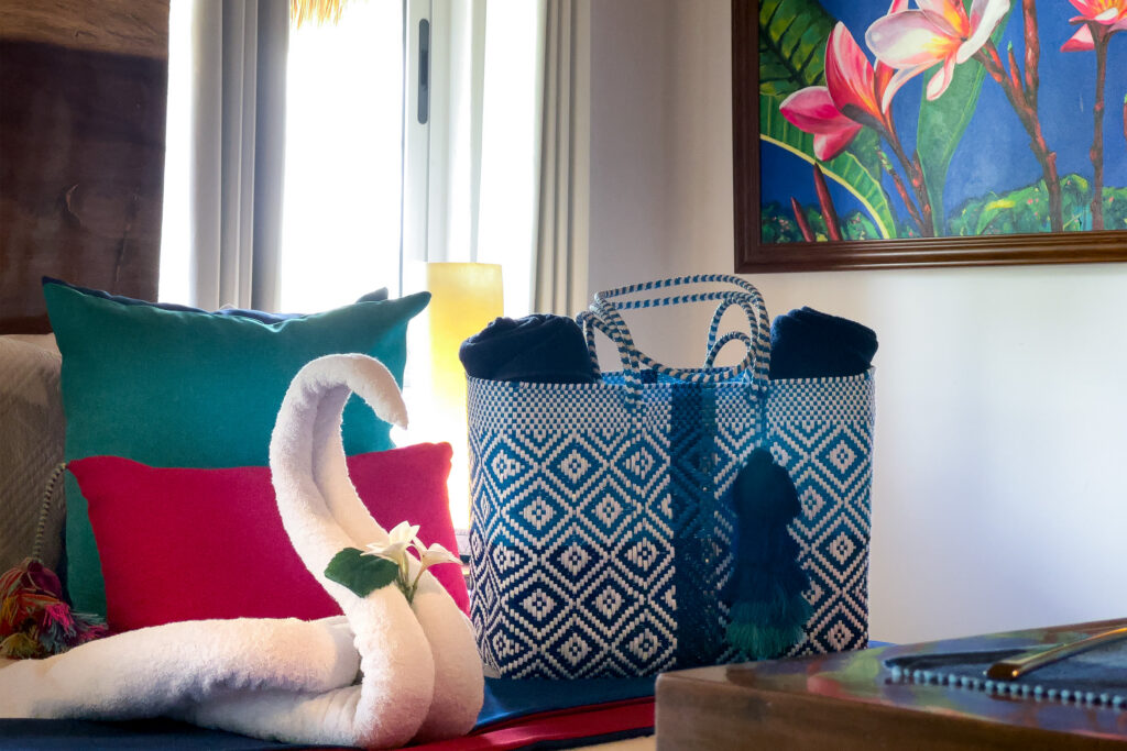 White towel swan beside a blue woven bag and bright pillows, with tropical artwork on the wall.