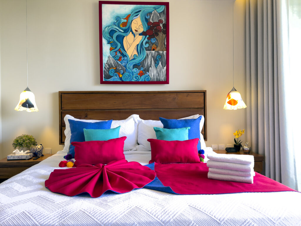 White bed with layered colorful pillows and a red throw beneath framed artwork and pendant lamps.