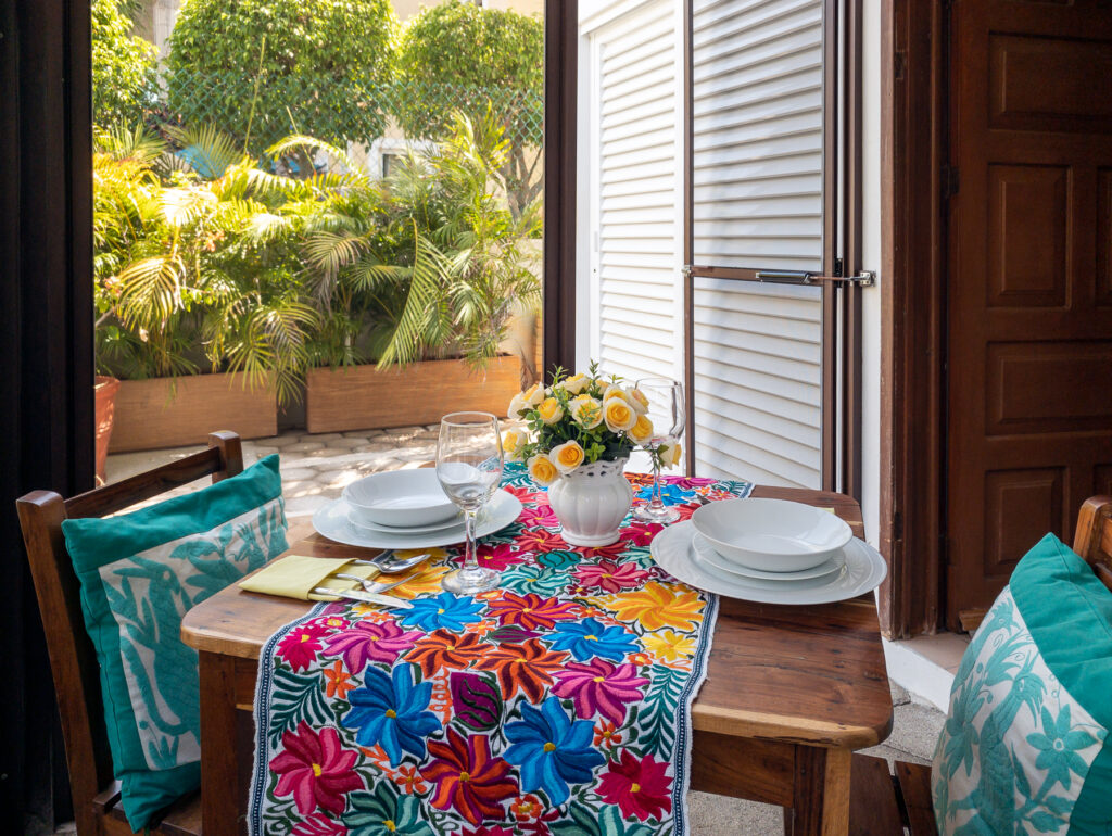Wooden patio table set with plates, glasses, and flowers over a bright floral runner.