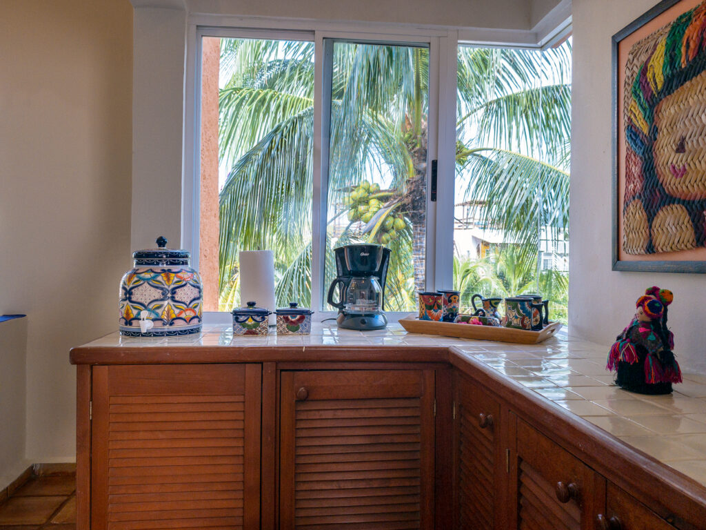 Coffee maker, cups, and decorated jar on a wooden counter beneath a window overlooking palms.